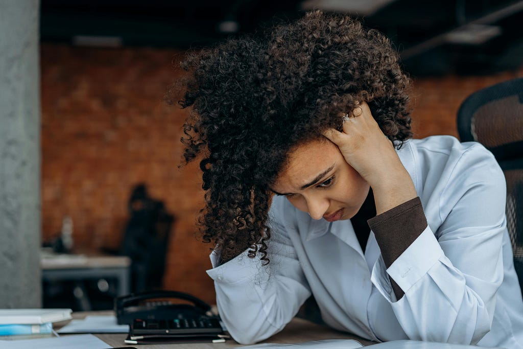 A frustrated black woman is sitting at her desk. Her hands are in her hair and she was a worried look on her face. She’s wearing a long sleeved button down baby blue colored shirt with a dark brown shirt beneath.