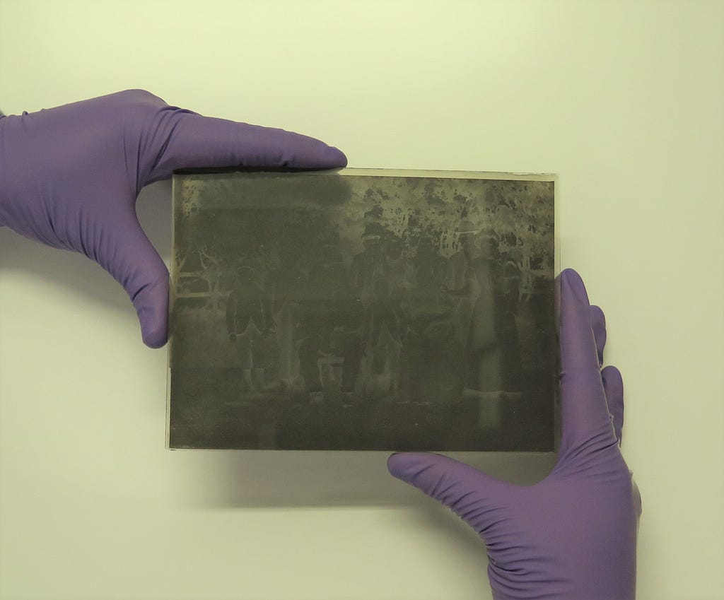 A conservator wearing purple nitrile gloves holds a glass plate negative on alternate corners by the edges by placing the thumb and forefingers in an L shape.
