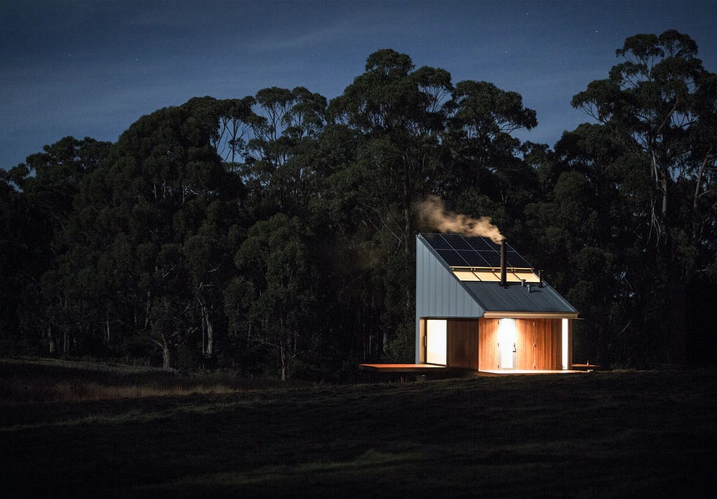 A small house with a tall, pitched roof is lit up in a dark, forest environment.