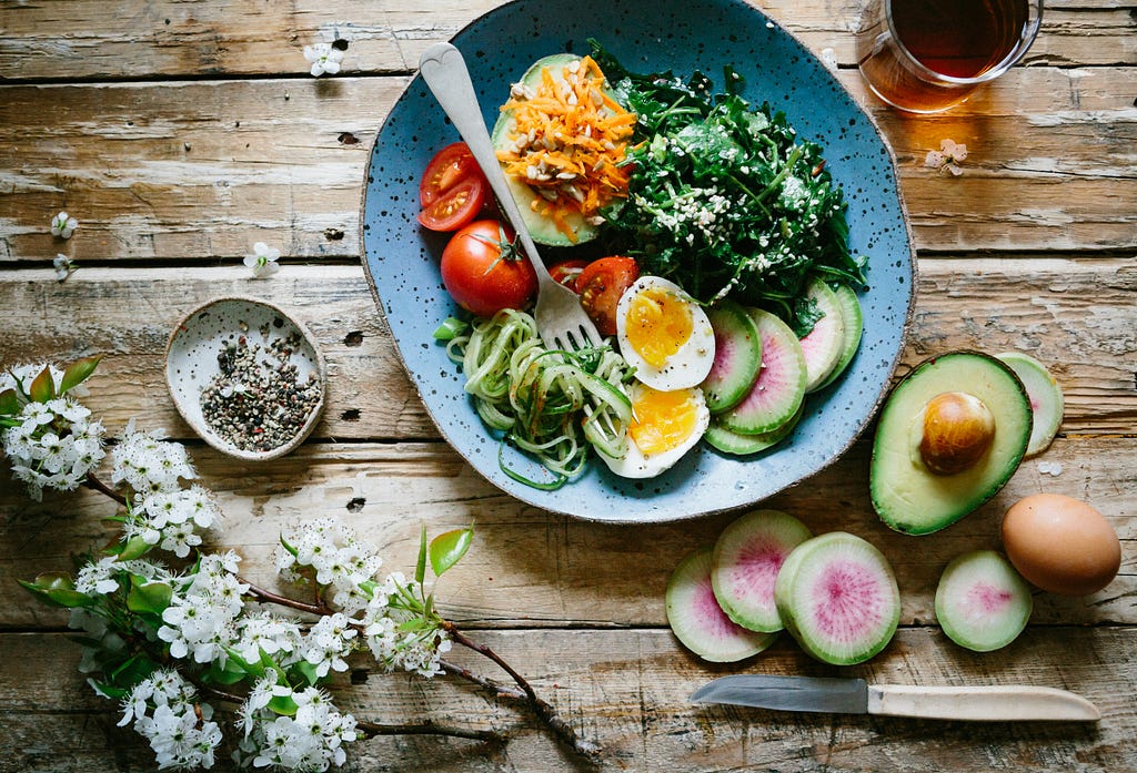 This image showcases a wholesome meal on a rustic wooden table, featuring a colorful array of fresh ingredients like tomatoes, cucumber spirals, radish slices, avocado, and a salad sprinkled with sesame seeds. A soft-boiled egg, a spice bowl, a glass of tea, and white flowers add to the natural and vibrant setting.
