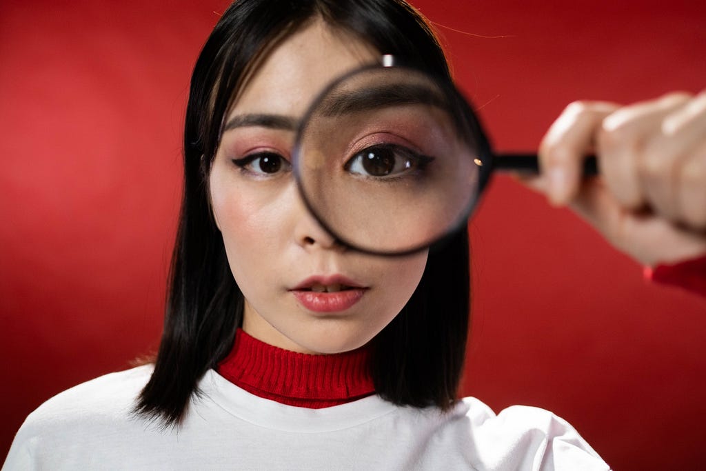 Picture of a woman with shoulder length dark hair standing in front of a red background. She is looking at you through a magnifying glass.