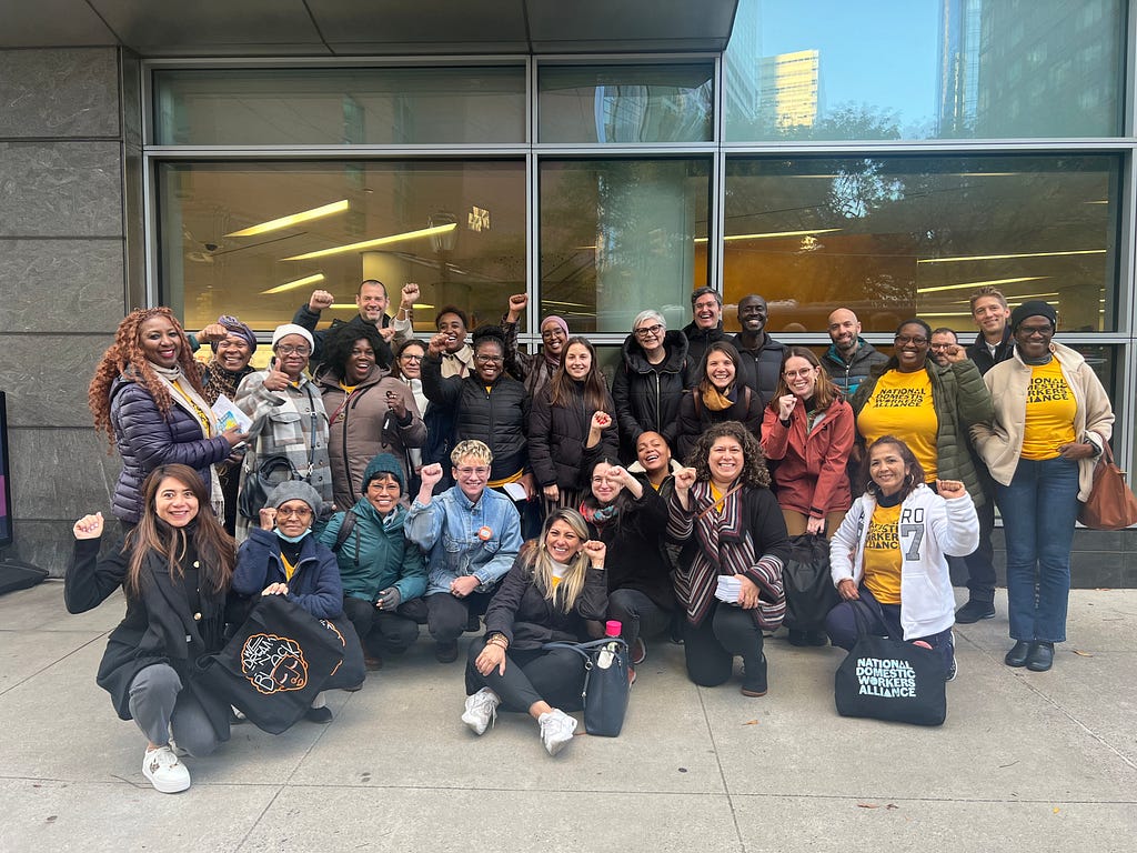 A group of people stand smiling in front of a public library in New York. Some are wearing t-shirts from the National Domestic Workers Alliance.