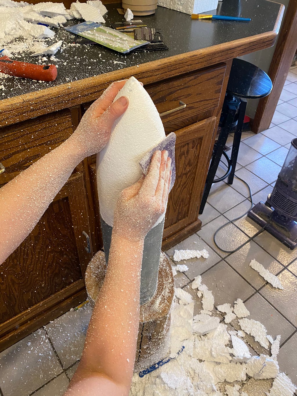 A piece of styrofoam being sanded into the shape of a rocket ship, leaving bits of foam all over the kitchen counters and floor.