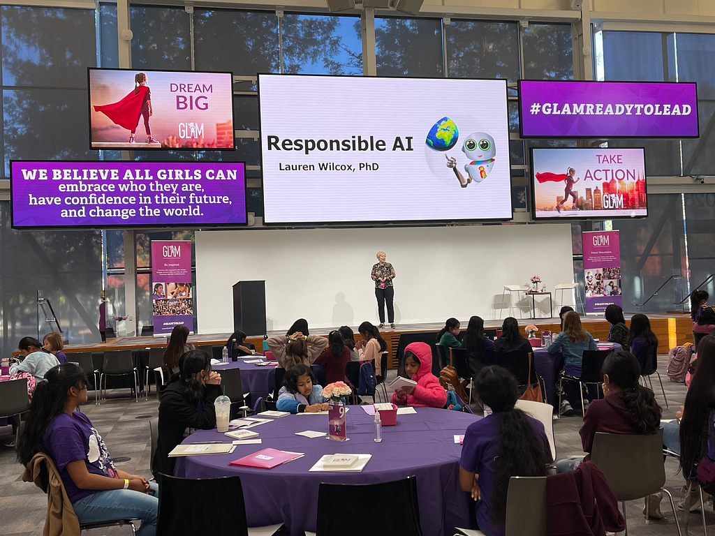 Standing on stage speaking to a large room of generation alpha attendees, who sit at round tables. The title of the slide shown above me is “Responsible AI”, with Girls Leadership Academy Meetup signage around it, including signs to Dream Big, and Take Action