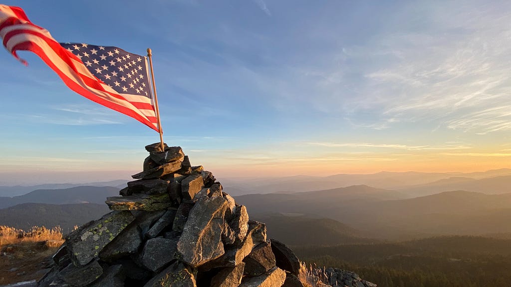 An American flag blows in the wind, its pole embedded in a shrine of gray rocks at the top of a mountain overlooking the hazy, rolling horizon under a blue sky.