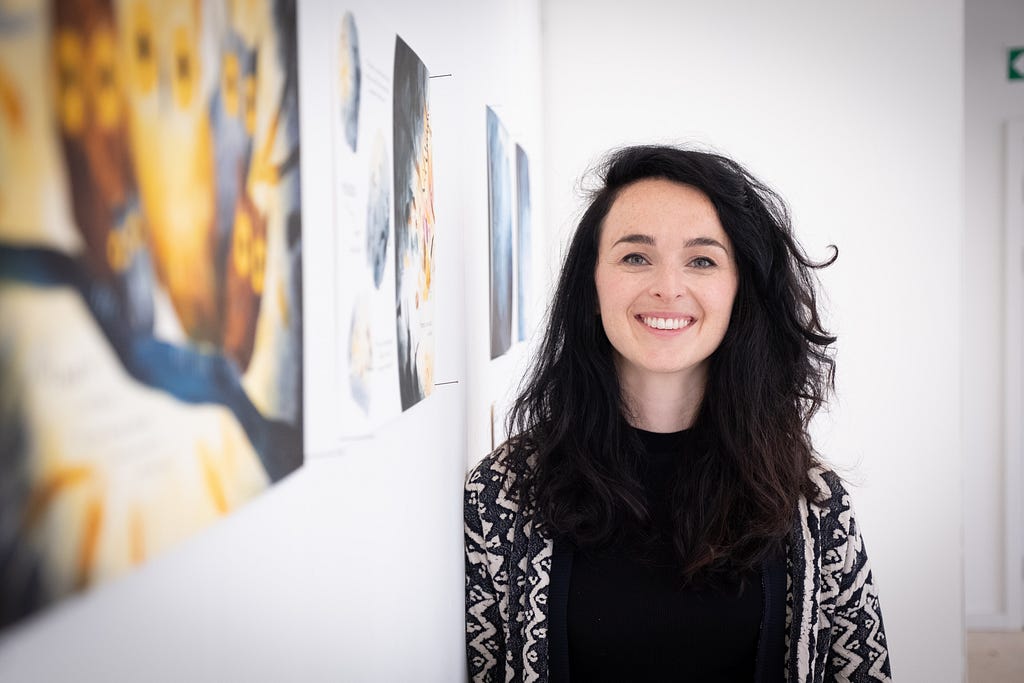 Photo of the writer and illustrator Helen Kellock standing next to a gallery wall displaying their illustrations, smiling cheerfully.