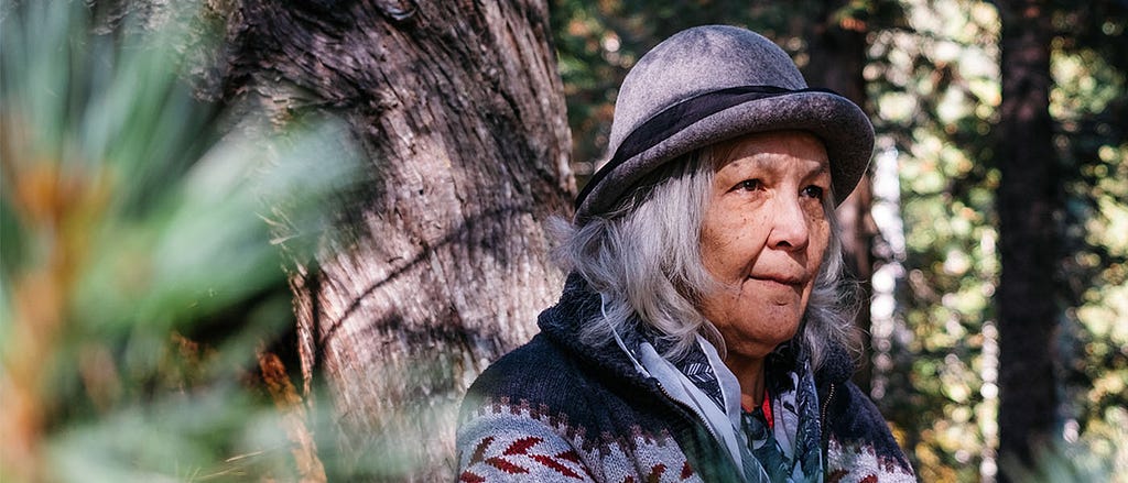 Sinixt Matriarch Marilyn James is shown in front of a tree, surrounded by forest, wearing a grey hat and patterned sweater.