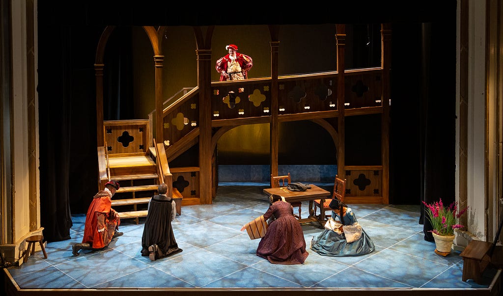 A man in red and gold 16th century clothes stands on a wooden platform looks down at four figures bowing deeply in robes and full skirts.