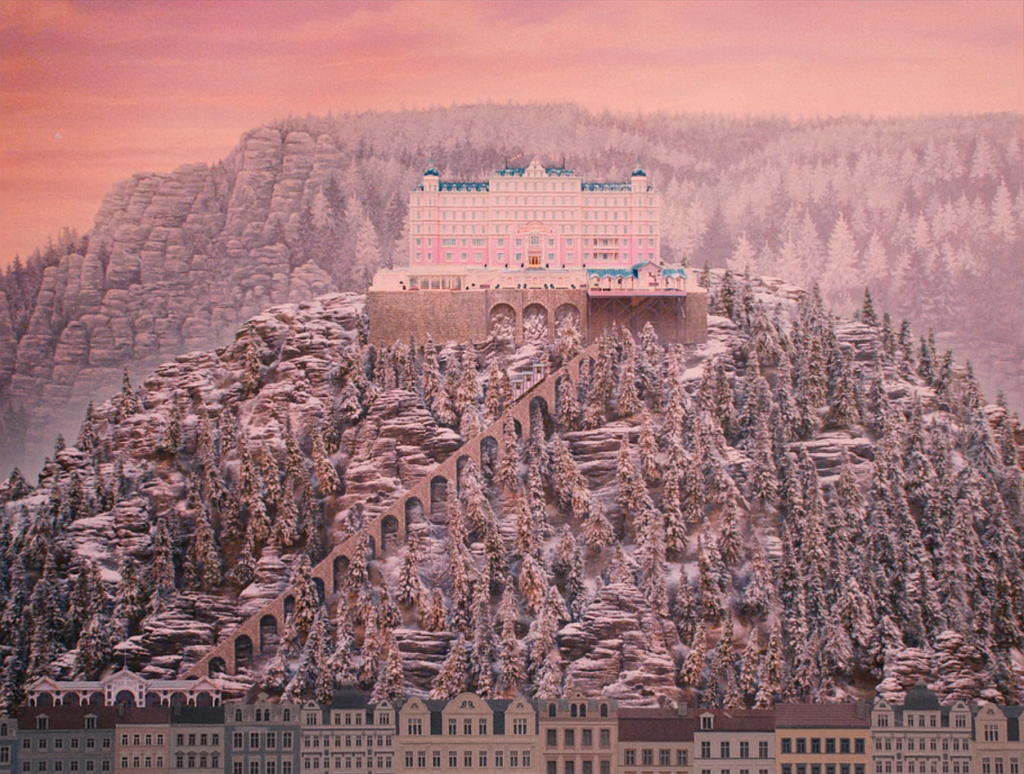 A lavish, pink hotel sits atop a tall, snowy mountain against a pink-er sky and tree-crested alps