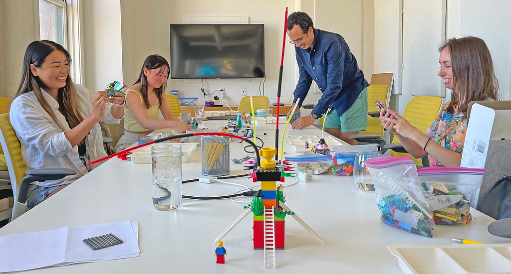 Four members of the design team participating in the building portion of a session, attaching pieces, holding models up, taking photos, and all smiling. The table they are working on is messy but colourful as it’s covered with drinks, pens, other models, and lots of LEGO bricks.