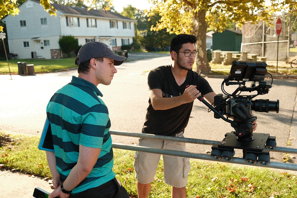 A camera moves along a dolly operated by one person with a second looking on.