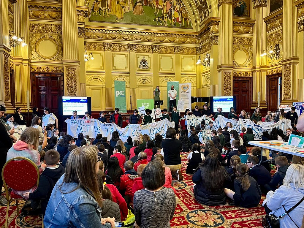 A diverse crowd mainly seated on the floor in a spacious room as a group of school kids hold up a banner.