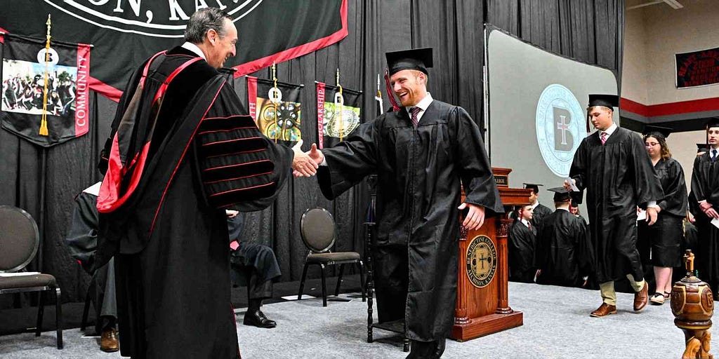 Benedictine College President Stephen D. Minnis on a stage shakes hands with a 2024 male graduate of the Catholic college in Atchison, KS.