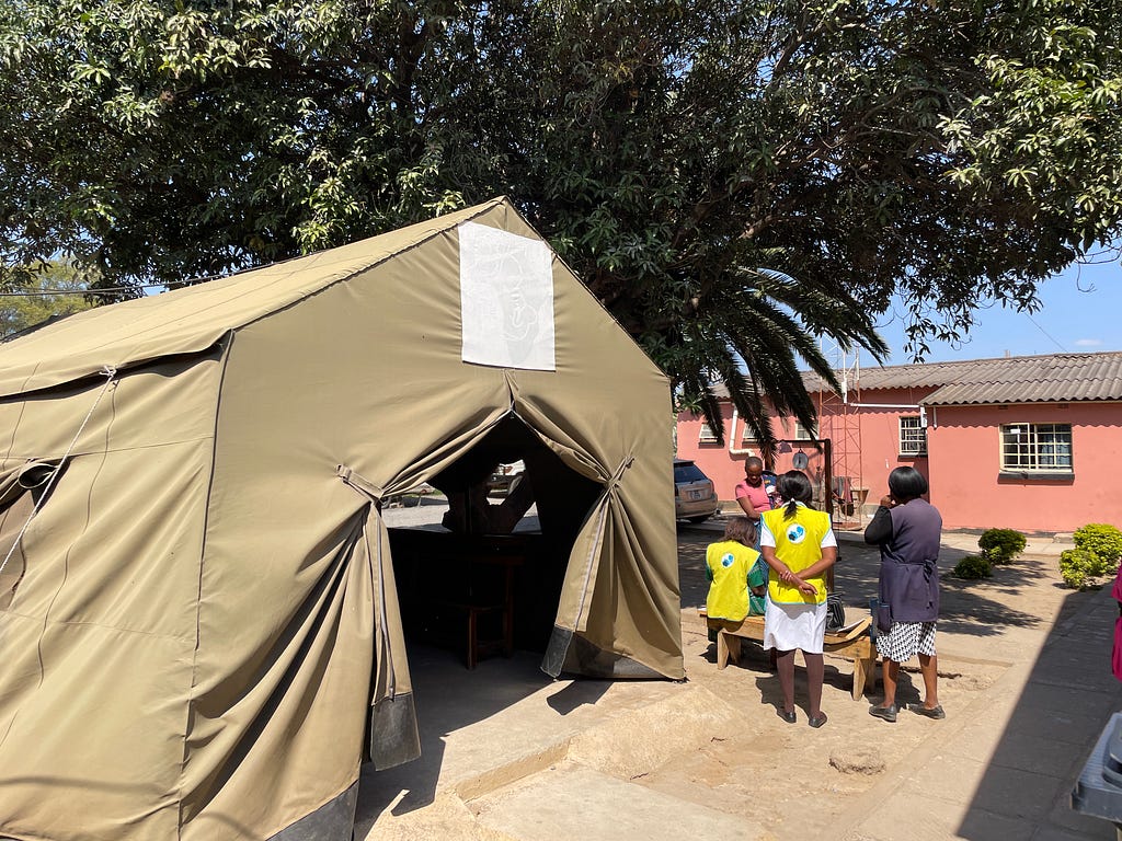 Multiple women at various stages of pregnancy, and health care workers, are walking in the courtyard of Bauleni’s Urban Clinic, next to a beige tent where counseling groups are held.