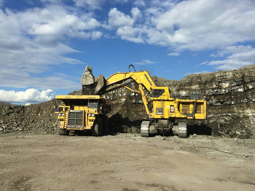 One yellow digger pouring rock and dirt into a large yellow lorry in an open air mine.