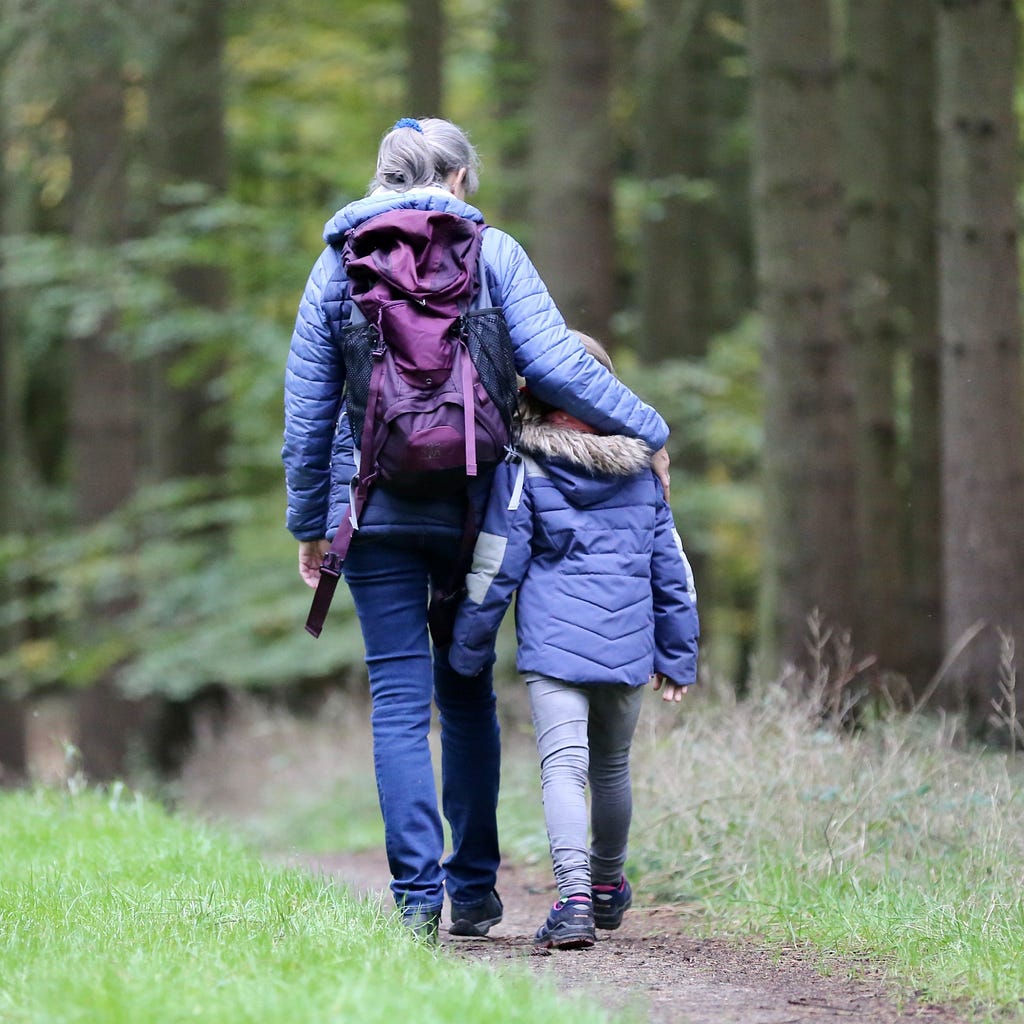 An older woman walking in the woods with a young girl, the older woman has an arm around the girl’s shoulders