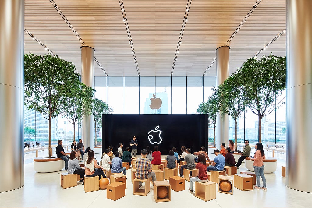 n einem großen Raum sitzen mehrere Menschen auf Hockern vor einem großen Bildschirm, auf dem das Apple-Logo zu sehen ist.