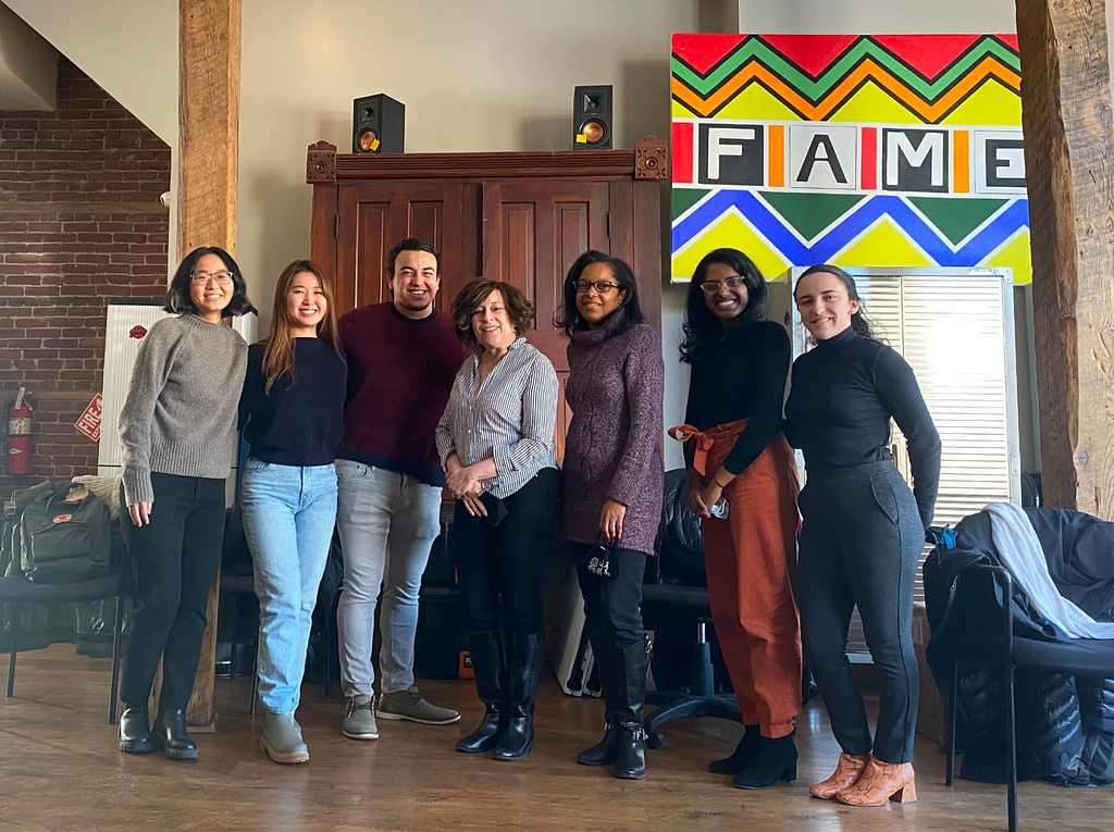 Seven people from the CMU Design Team and FAME staff stand together, smiling, in a spacious, cozy office room decorated with a colorful painting of the FAME logo.