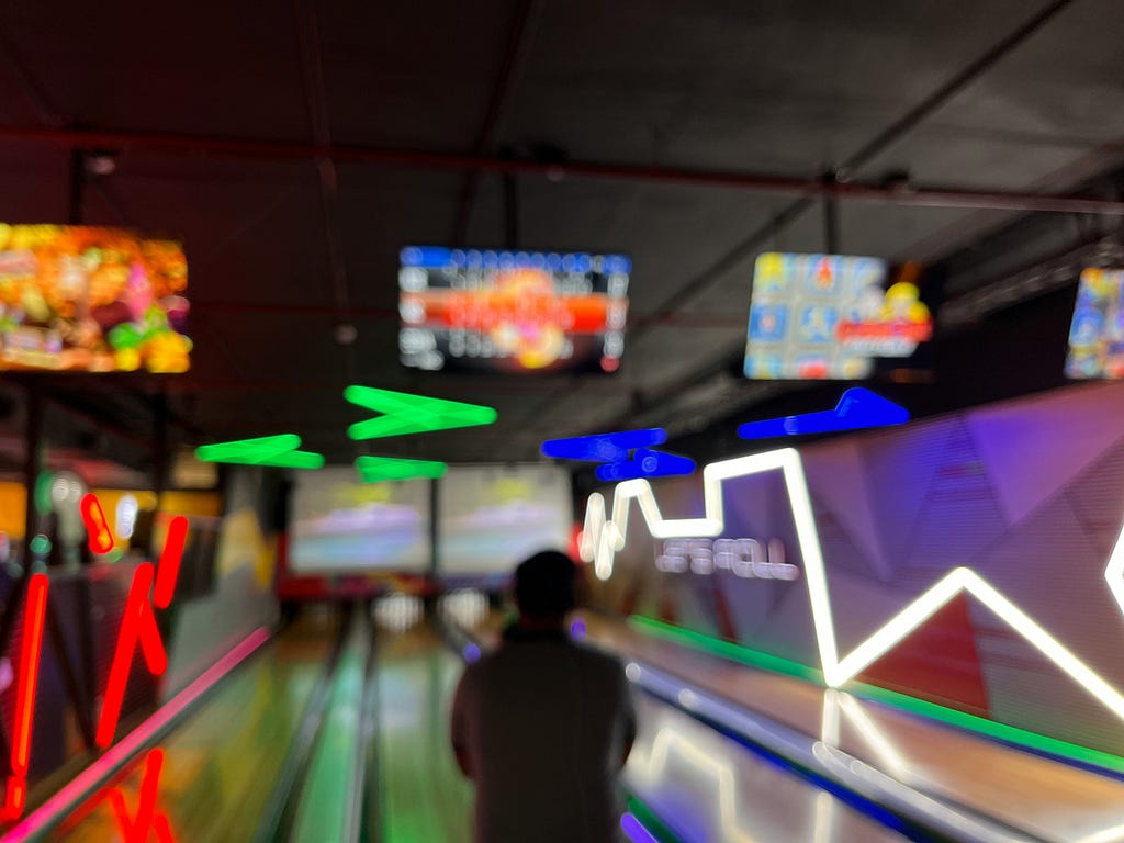 A person, how hands are hidden in front of them, stands just before an open lane at a bowling alley. All pins are set for a fresh take at the bowl. Neon lights are all around, the room feels dark and cool. It is ready to be played.