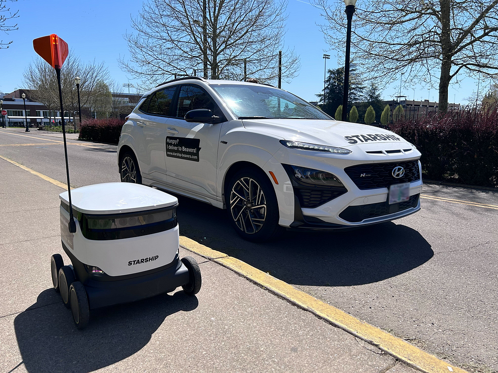 Image of a Starship Robot next to a white car with decals that read “Starship” and “Hungry? I deliver to Badgers” to make the white car look like a Starship Delivery Robot