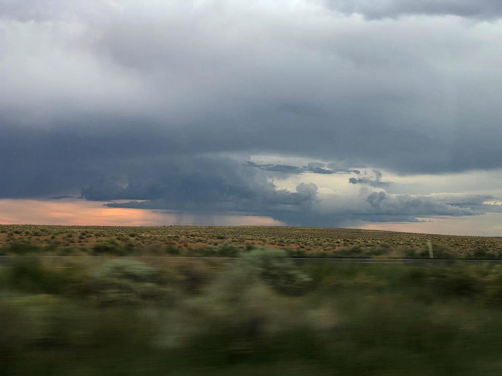 Photo from a moving car,  foreground is blurred, and beyond fields of sage brush, there is a sunset and two rain storms.
