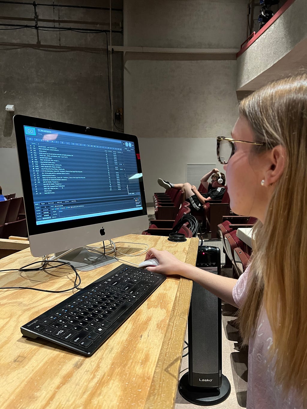 A theater student sits in front of a computer displaying a sound designing software at a wooden desk.