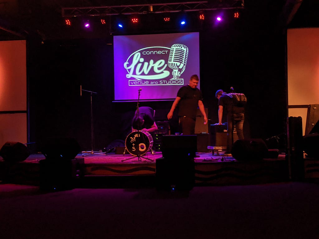 Three people set up equipment on a small stage. There is a projection on a screen above that reads, “Connect Live.”