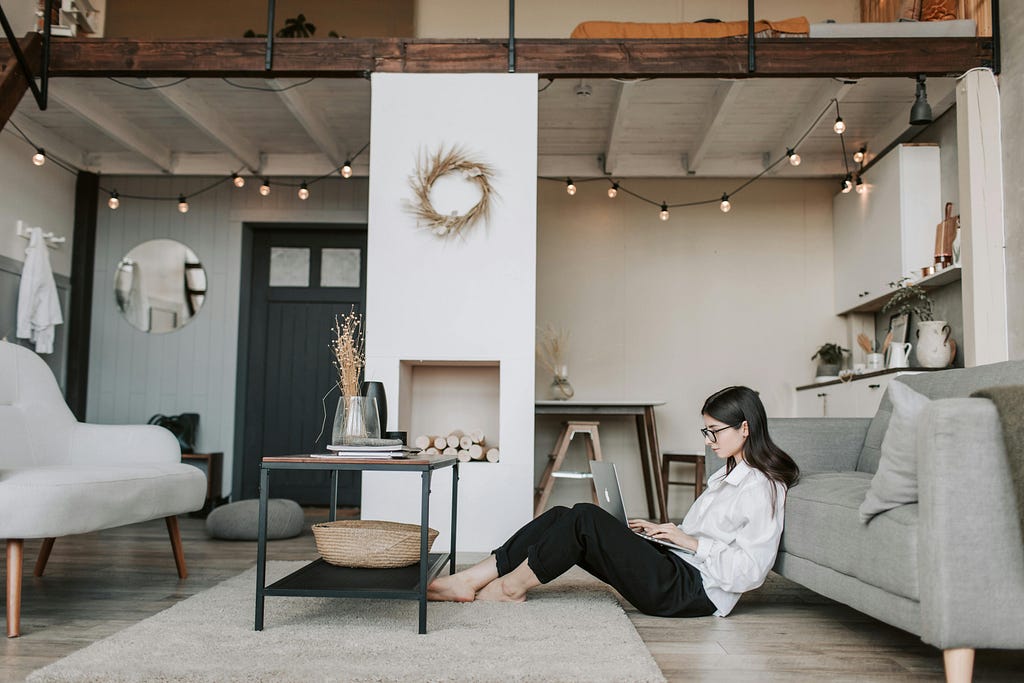 A woman in her 20s sitting on the floor at her white-beige, contemporary designed apartment, working on laptop.