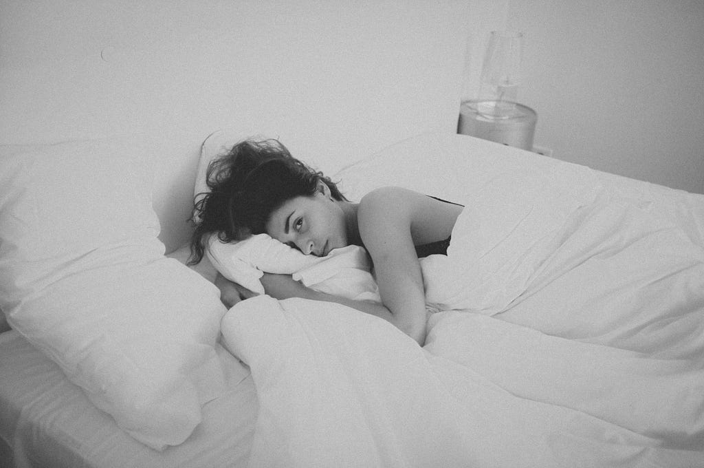 A greyscale photo where a woman lies in an all white bed wrapped in blankets and grasping the pillow under her head.