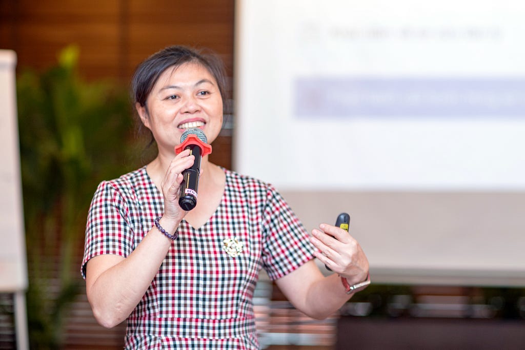 A woman giving a presentation holds a microphone in one hand a gesture with her other hand which is grasping a presentation clicker. A blurred image of a presentation appears behind her on a large display.