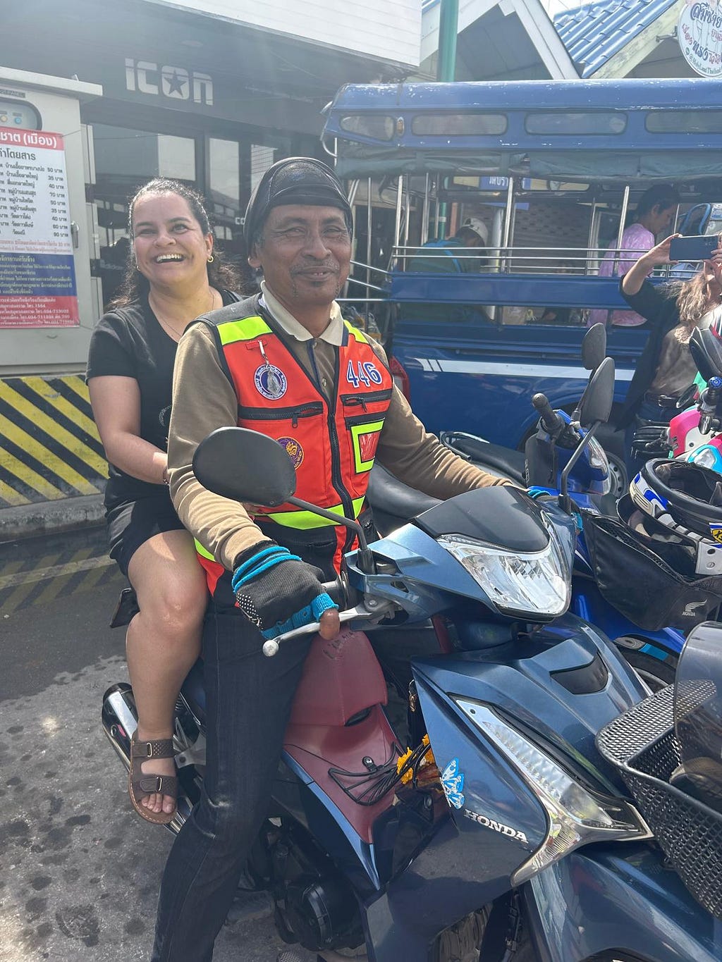 Un hombre y una mujer comparten una motocicleta en Tailandia. Ambos sonríen a la cámara. El hombre porta un chaleco de seguridad.
