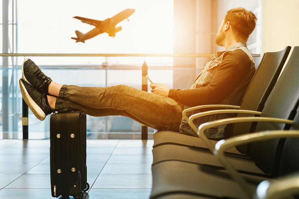 A man sits in an airport with his feet resting on his luggage, watching a jet outside take off.