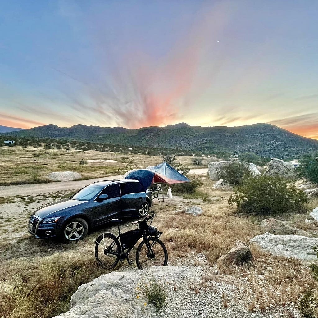 Car, bicycle and tent in the foreground of a desert, with the sunset in the mountains in the background.