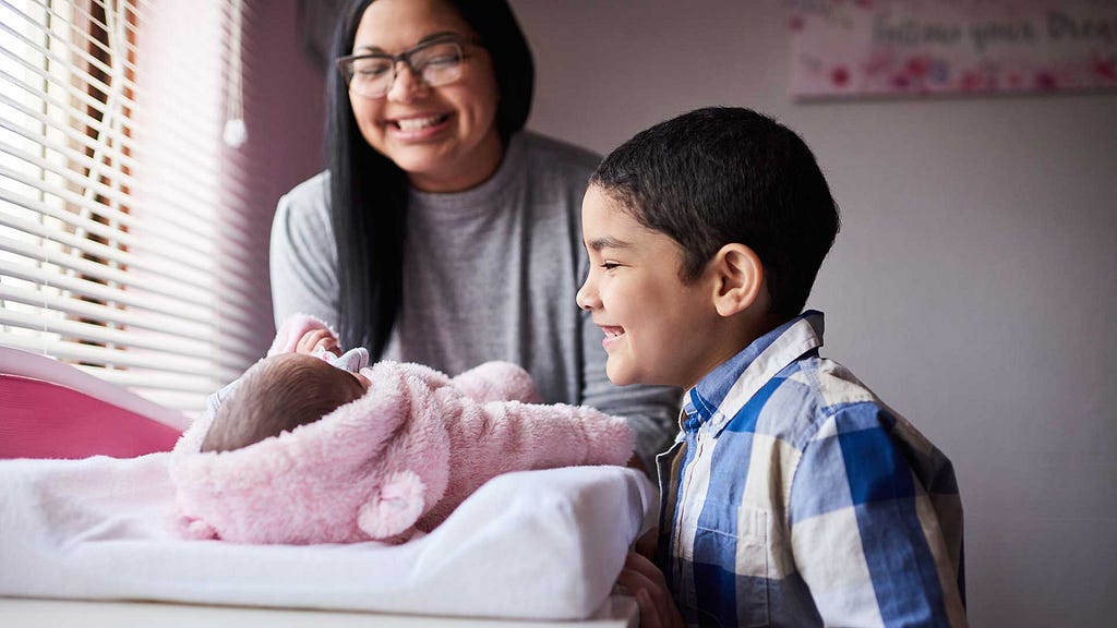 Mom and young boy smile at baby on a changing table.