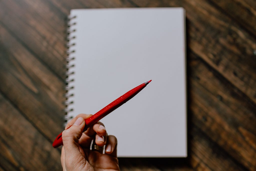 A left hand holds a red pen poised over a notebook on a wooden desk.