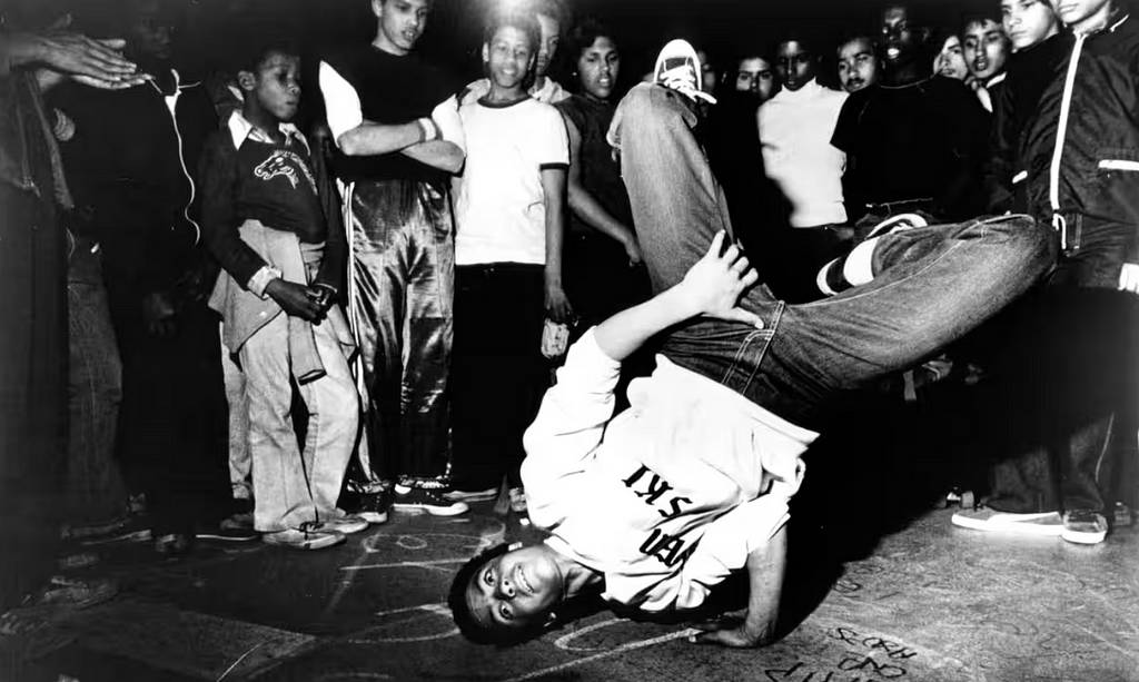 A breakdancer is doing a freeze in a cypher while the crowd around looks at him in glee.