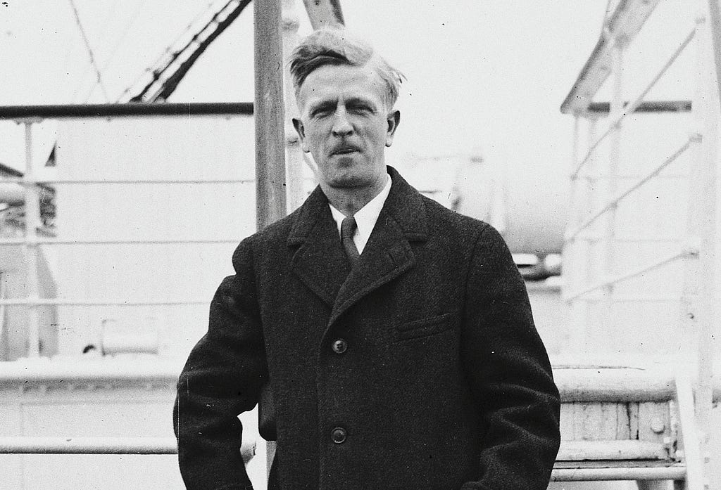 The same man as in the earlier photograph stands in front of stairs on the deck of a ship. His hair is windblown. He is wearing a black overcoat, a tie, and a white shirt.