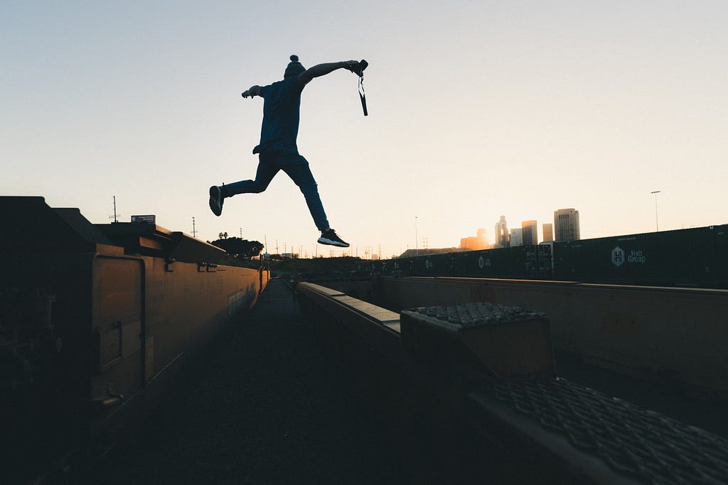 Man holding camera jumps from the top of a train to a nearby platform as the sun sets.
