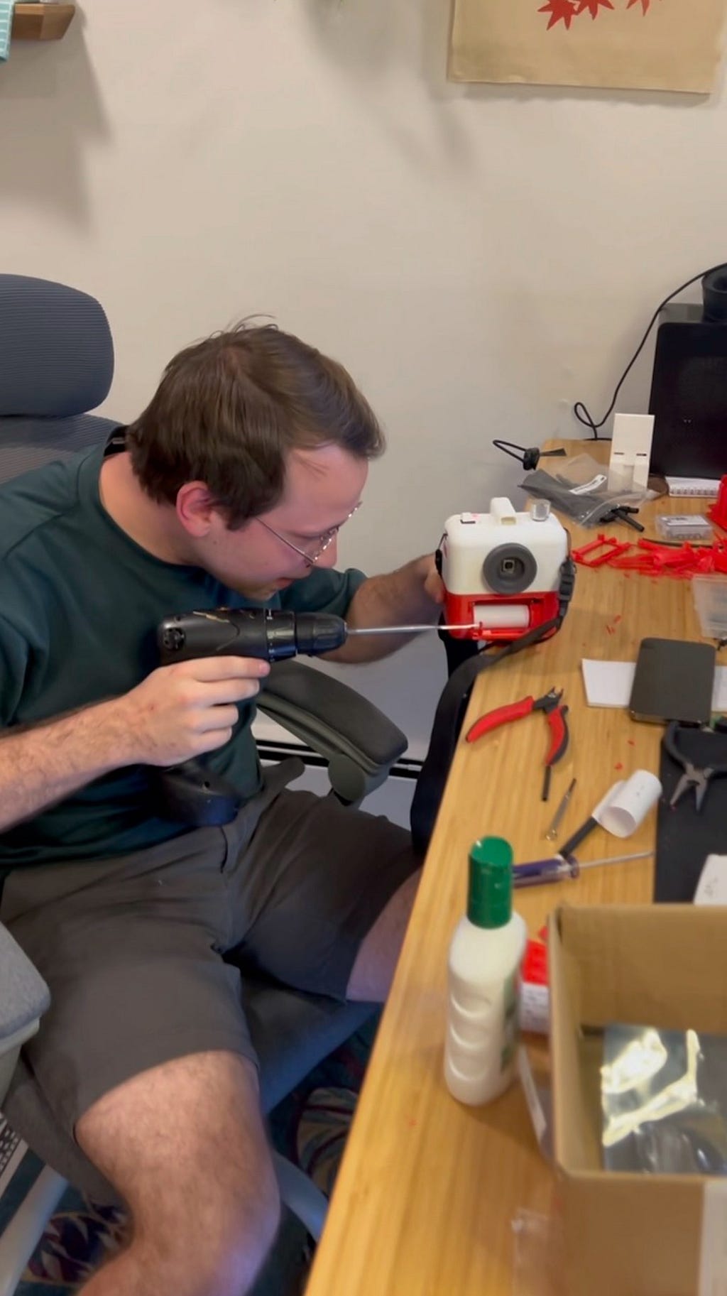 Ryan sits at a desk holding a power tool, which is being used to construct Poetry Camera.