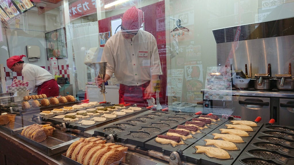 Through a glass screen, a taiyaki-maker adds some finishing touches to a large tray of the stuff.