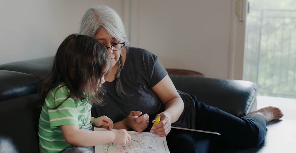 A mother and her child sit on a couch, coloring together.