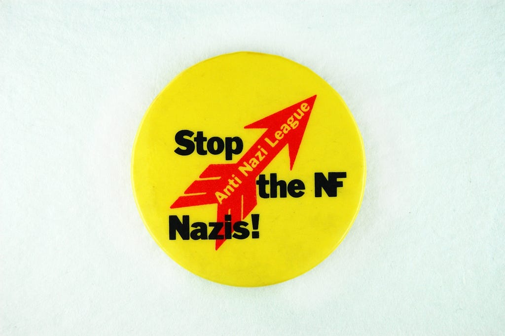 Plastic protest badge. Red and black design on yellow circular badge, with text reading Anti Nazi League; Stop the NF Nazis!