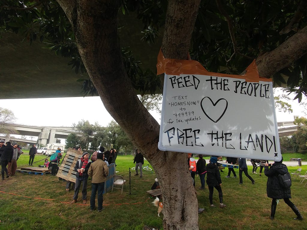 A group of people gathered in a park building tiny homes. A sign reads “Feed the People Free the Land!”