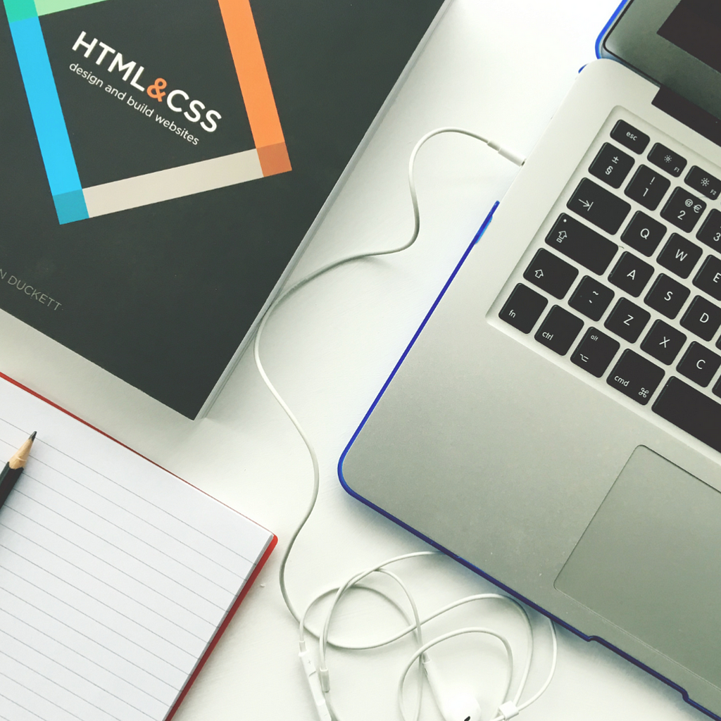 A photo showing a notepad, pencil, a book about web designing and a laptop. All with what resembles white earphone wire in the middle of the table.