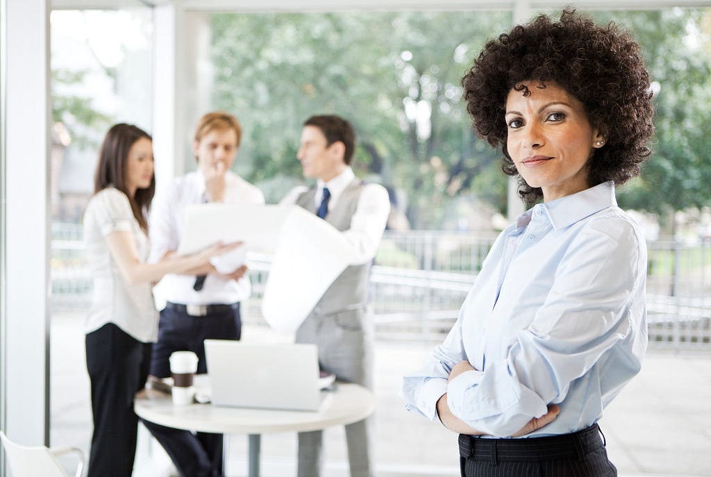 A woman looking in the camera with three of her colleagues behind her having a discussion