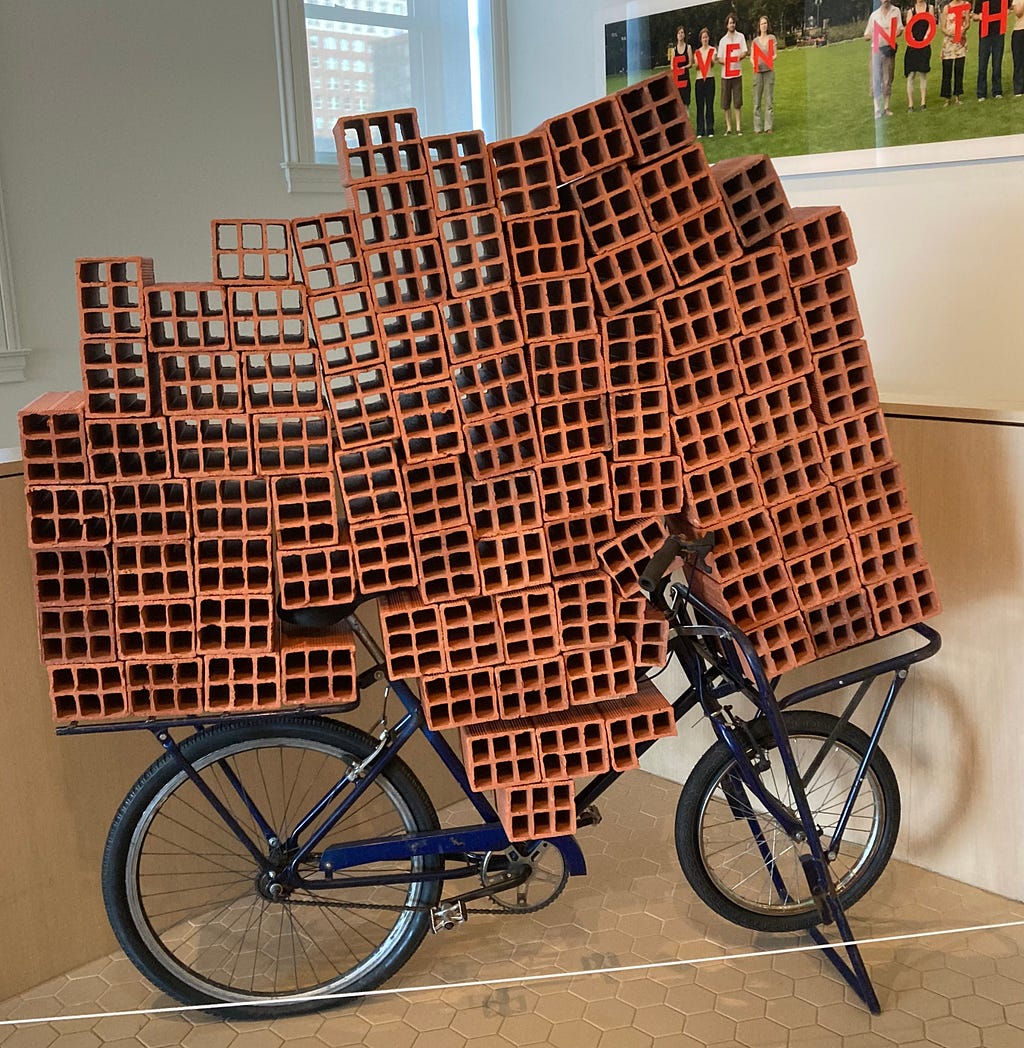 Dozens of large red clay mud bricks loaded on a black metal bicycle in an art installation of 21st century art. The bricks seemingly overwhelm the bike, which is still, however, standing.