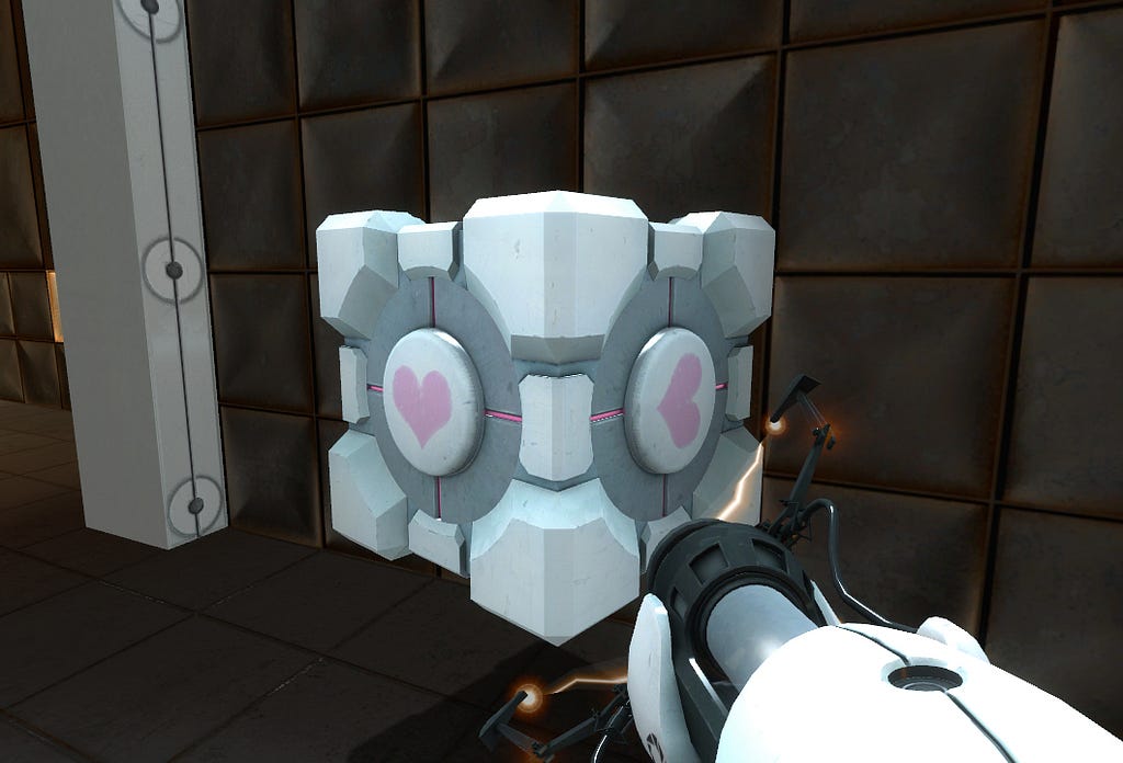 A picture of the companion cube from Portal. It is a white-and-grey metal cube suspended in midair, with a pink heart painted on each of its sides. The portal gun, a white-and-black futuristic contraption, protrudes from outside the bottom right corner of the image, holding the cube. There is a wall of black metal in the background.