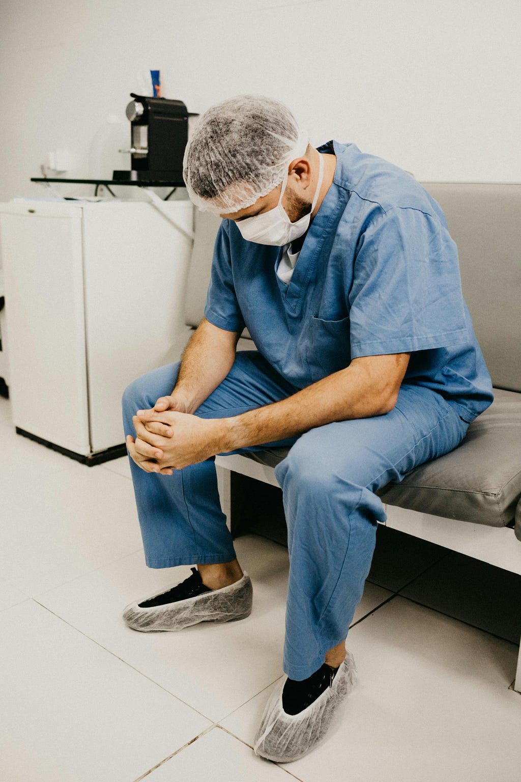 A doctor in scrubs sits on a couch with his head down in an on-call room. The man looks tired.