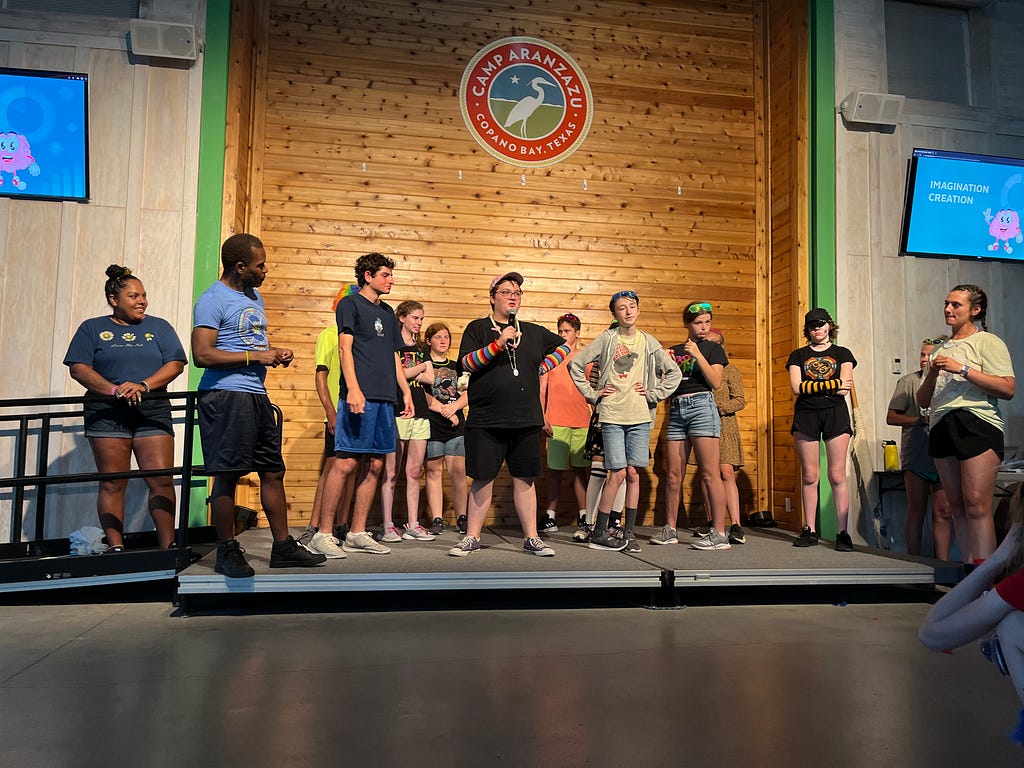 campers in imagination coalition gather around to show off their skills on stage at the HOST showcase in the dining hall. One camper has the mic and is speaking to the crowd while the rest of the group supports him on stage.