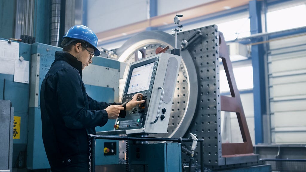 A man changes the setting for a plastic injection molding machine in an off-shore or domestic manufacturing facility while wearing a jumpsuit and plastic hardhat.